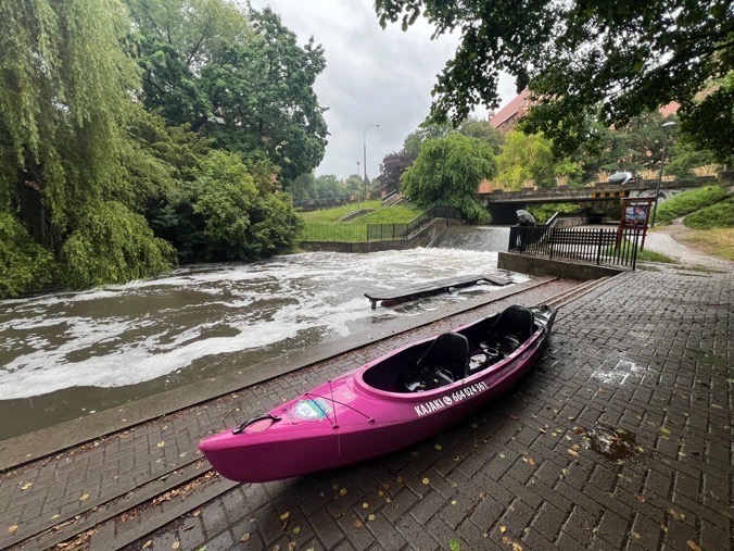 Różowy kajak dwuosobowy z numerem telefonu na brzegu rzeki z bystrzem i zielenią w tle, w pochmurny dzień.