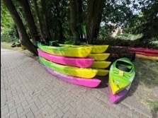 Stos kolorowych kajaków (żółto-zielonych i różowo-fioletowych) ułożonych na brukowanej ścieżce pod drzewami, czekających na wynajem w plenerze.