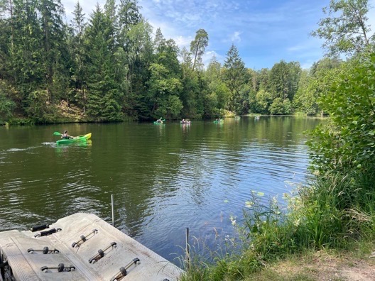 Spokojny krajobraz jeziora z kajakami, zielone drzewa w tle, pomost na pierwszym planie. Idealne otoczenie na imprezy plenerowe i relaks na wodzie.