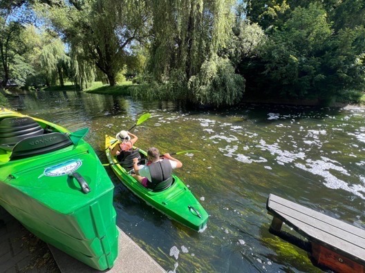 Dwie osoby płynące kajakami po rzece w słoneczny dzień. Jeden kajak zielony na brzegu, drugi na wodzie z parą w kamizelkach ratunkowych.