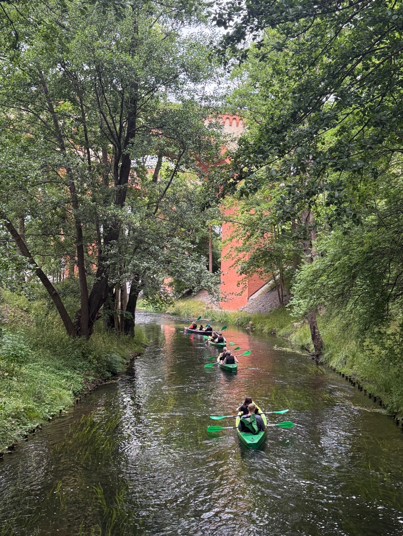 Spływ kajakowy malowniczą rzeką, z widokiem na ceglany most i bujną zieleń. Kajaki w jaskrawych kolorach. Aktywny wypoczynek na łonie natury.