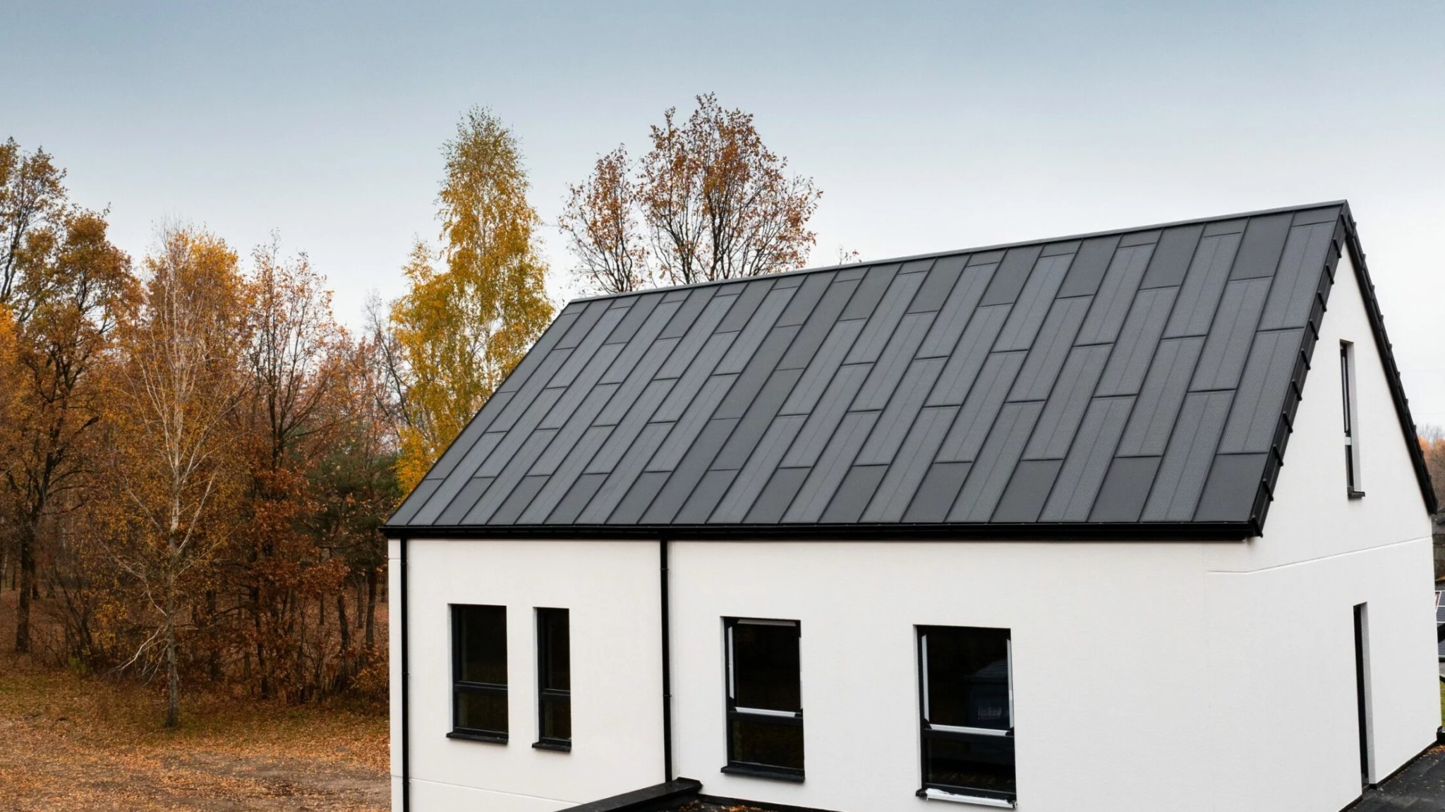 Biały dom z ciemnoszarym dachem panelowym na tle jesiennego krajobrazu. Minimalistyczna architektura, nowoczesne wykończenie.