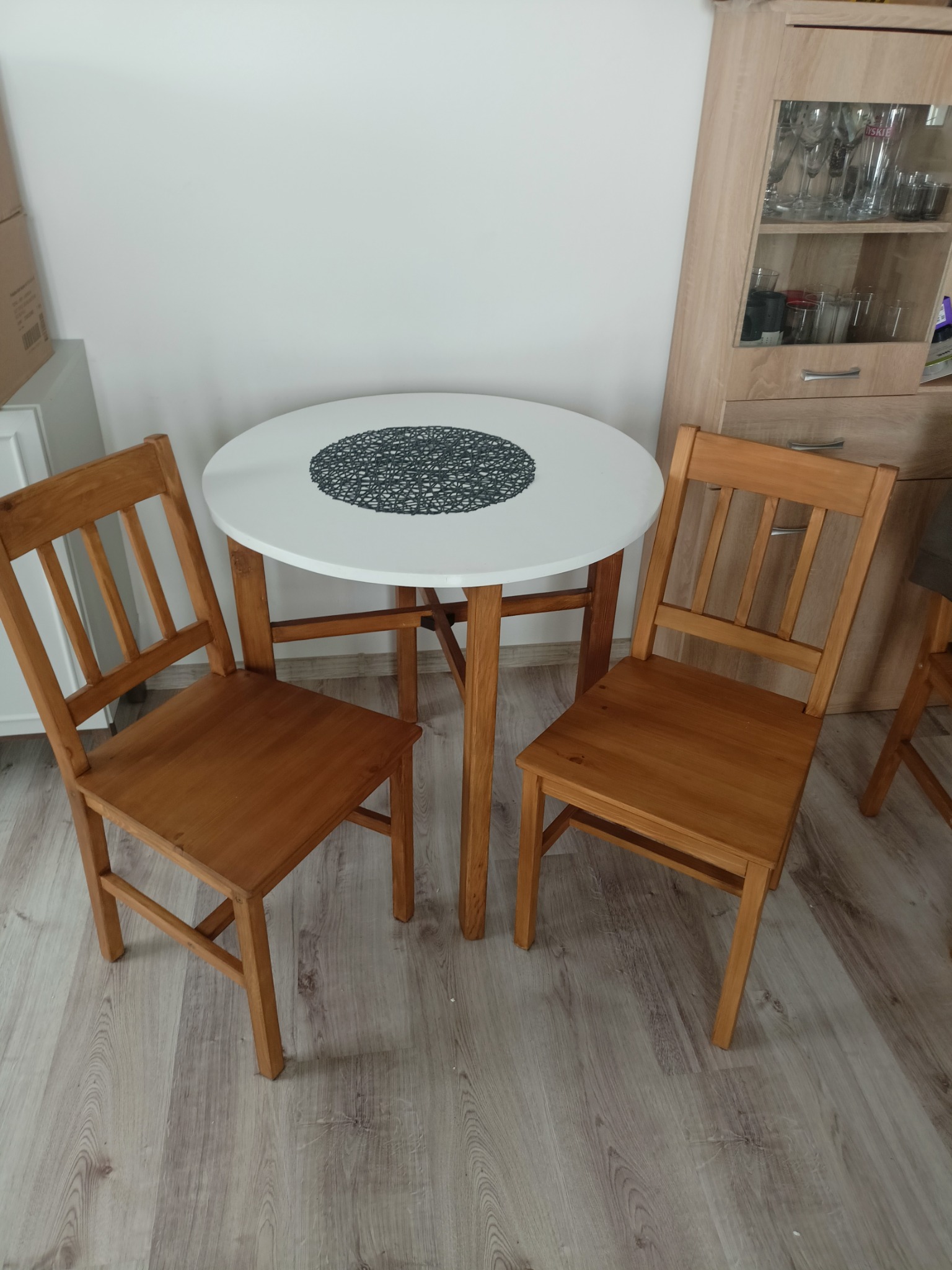 Drewniane krzesła i okrągły stół z białym blatem w jasnym wnętrzu. Na stole czarna podkładka, w tle szafka z przeszkleniami i kartony.