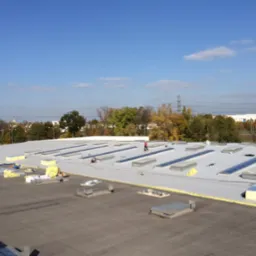 Jasny dach przemysłowy z licznymi świetlikami i dwoma pracownikami podczas montażu, w tle drzewa i słupy energetyczne, słoneczny dzień.