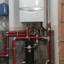 Biały piec Vaillant z czerwonymi rurami i czarnym filtrem, zamontowany na szarej ścianie z płytkami. Widoczny grzejnik i niebieski zbiornik wyrównawczy.