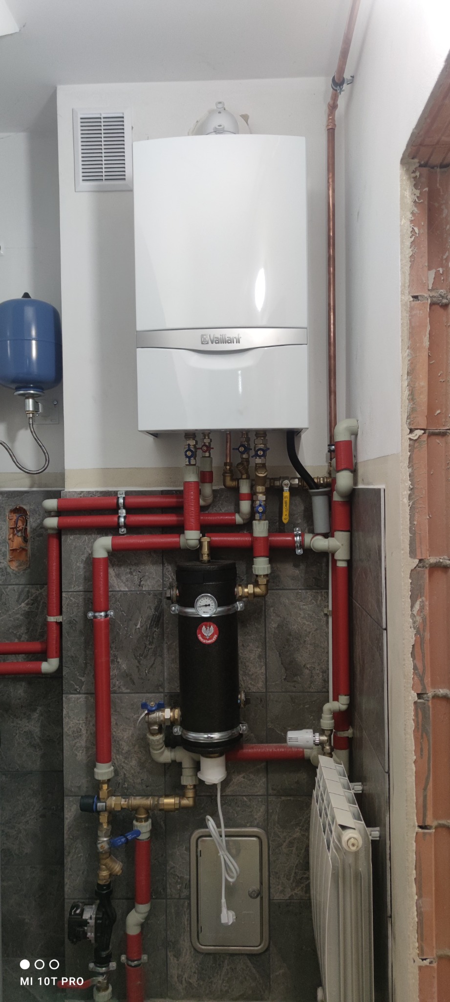 Biały piec Vaillant z czerwonymi rurami i czarnym filtrem, zamontowany na szarej ścianie z płytkami. Widoczny grzejnik i niebieski zbiornik wyrównawczy.