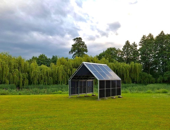 Szkieletowa konstrukcja z paneli solarnych na zielonym trawniku, w tle drzewa i pochmurne niebo. Nietypowa budowla z energią słoneczną w plenerze.
