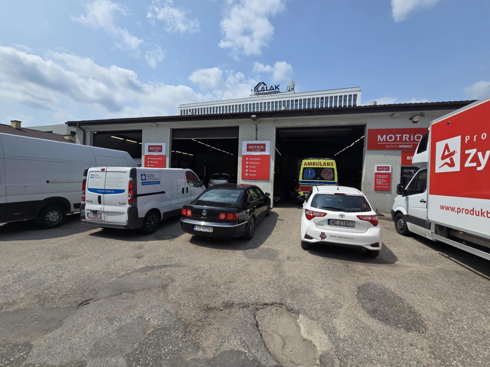 Warsztat samochodowy MOTRIO z zaparkowanymi pojazdami: karetka, Volkswagen Phaeton, Toyota Yaris. W tle budynek firmy LALAK Development.