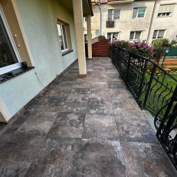 ProF-Bud - Ułożona, brązowa terakota na balkonie w Świnoujściu. Widok na budynek mieszkalny i ogród przez ozdobną, czarną balustradę. Estetyczne wykończenie podłogi.