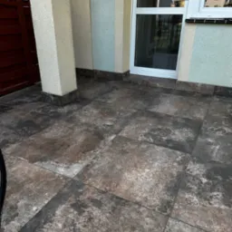 Wykończony taras z brązowo-szarych, kwadratowych płytek, kolumny elewacyjne i białe okno w tle.