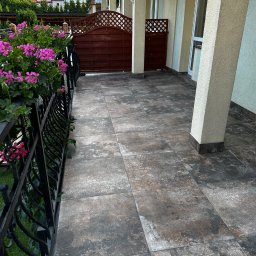 ProF-Bud - Wyłożony płytkami taras z kutą balustradą i kwiatami w donicach. Widok na dom z kolumnami i drewniane ogrodzenie.