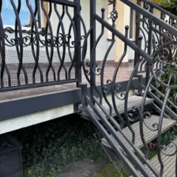 Metalowe schody zewnętrzne z ozdobnymi, kutymi balustradami, prowadzące do wejścia domu. Widoczne detale zdobień i ażurowe stopnie.