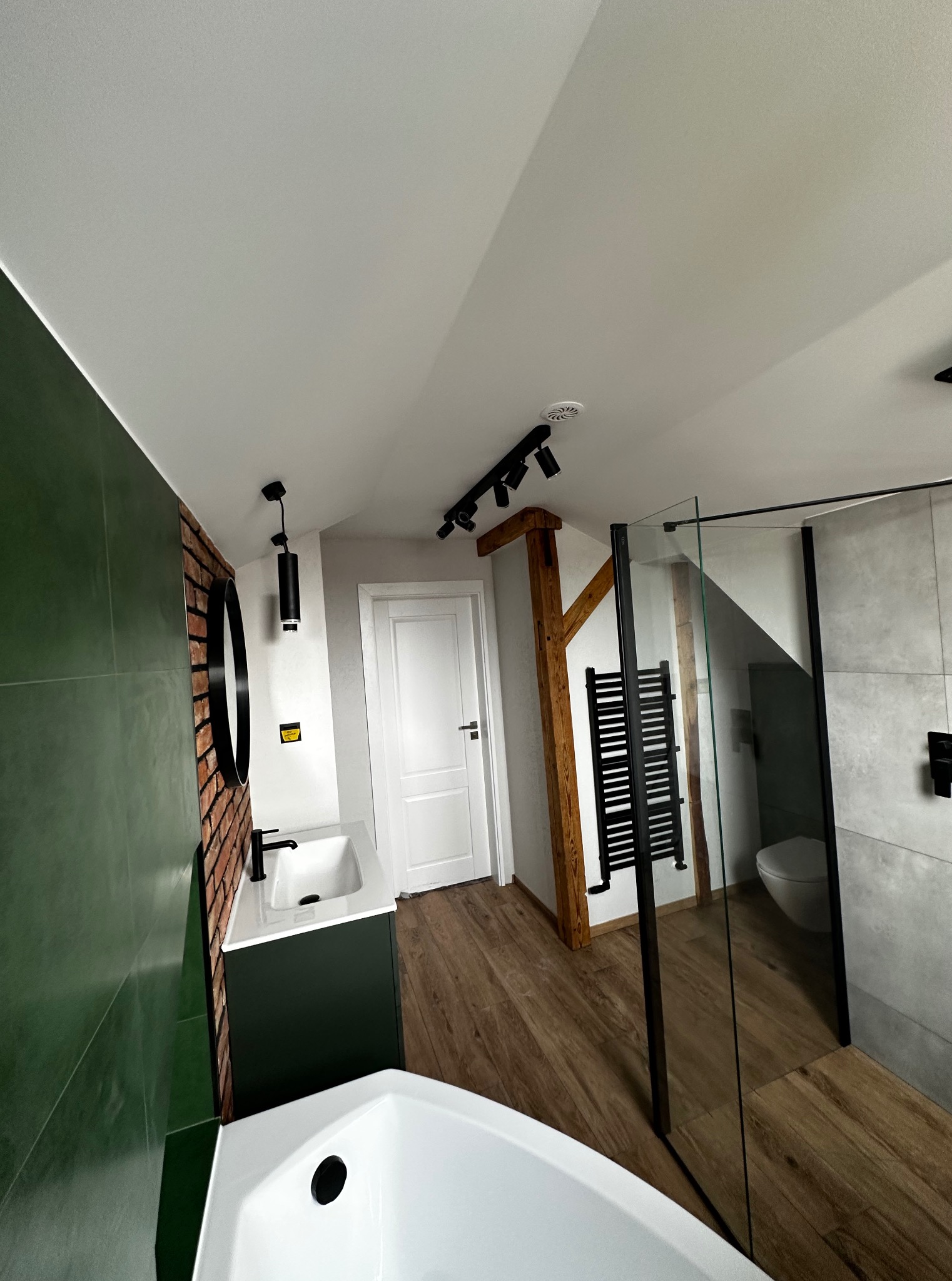 Nowoczesna łazienka na poddaszu z zielonymi płytkami, kabiną prysznicową ze szklaną ścianą i drewnianymi elementami konstrukcyjnymi. Widoczna umywalka i toaleta.