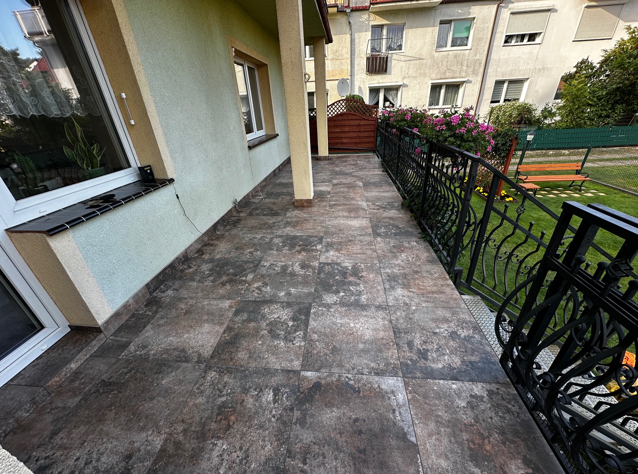 Wykończony balkon z brązowymi płytkami podłogowymi, czarną, ozdobną balustradą i widokiem na ogród z kwiatami. Słoneczny dzień.