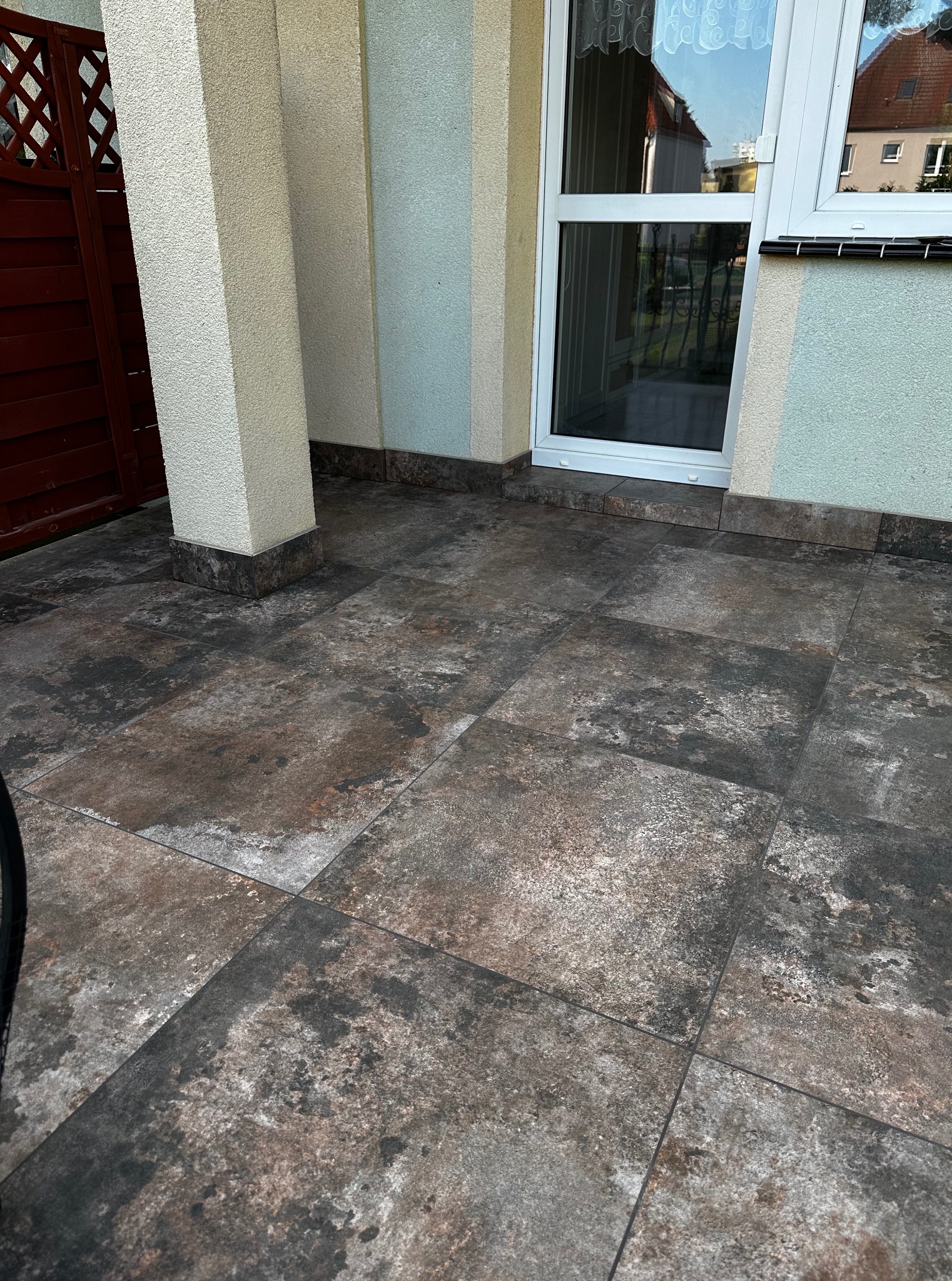 Brązowo-szare płytki podłogowe na zewnątrz budynku, widoczne kolumny i fragment okna z odbiciem nieba. Płytki ułożone równo, z widoczną fakturą.