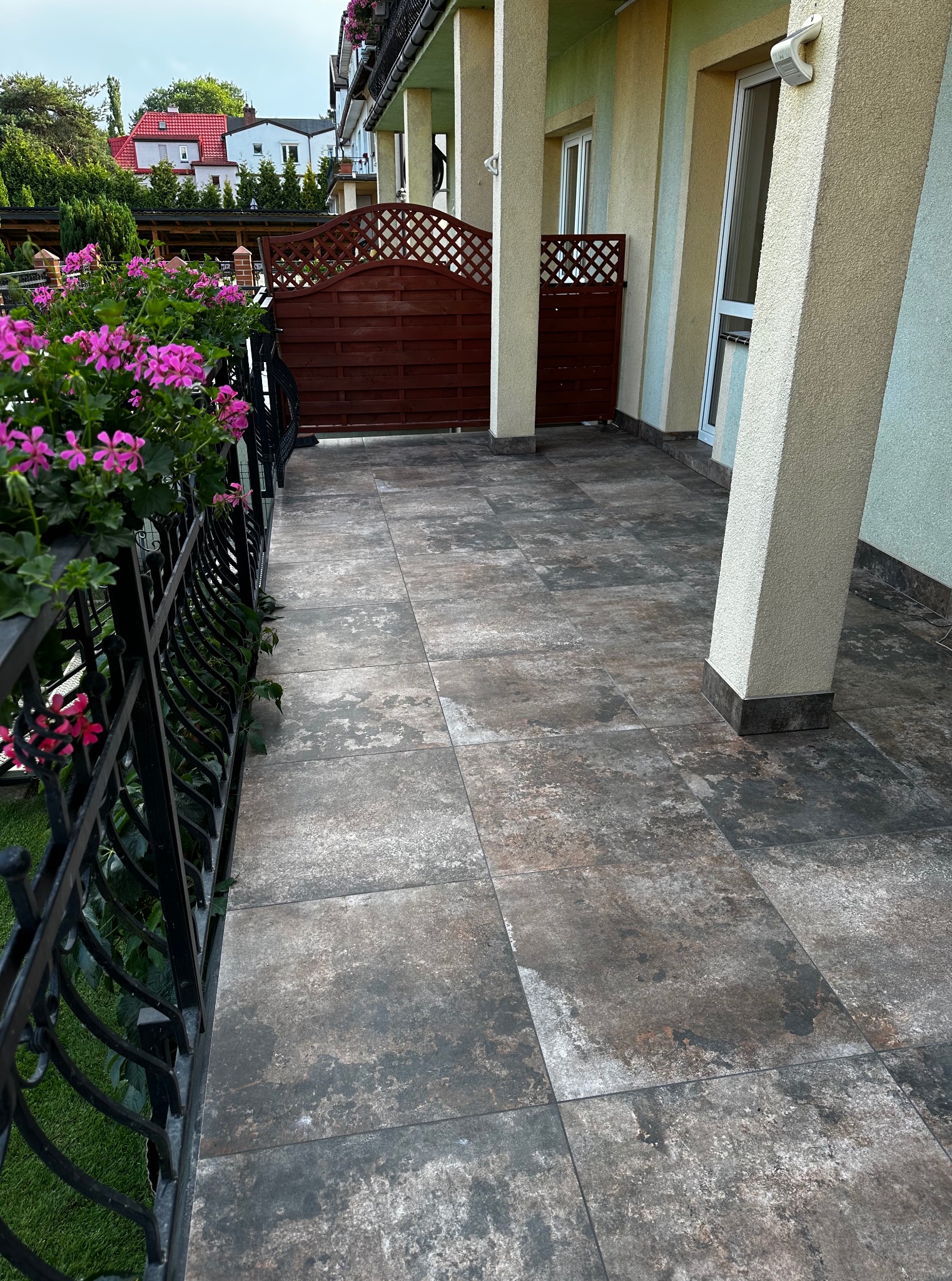 Wyłożony płytkami taras z kutą balustradą i kwiatami w donicach. Widok na dom z kolumnami i drewniane ogrodzenie.