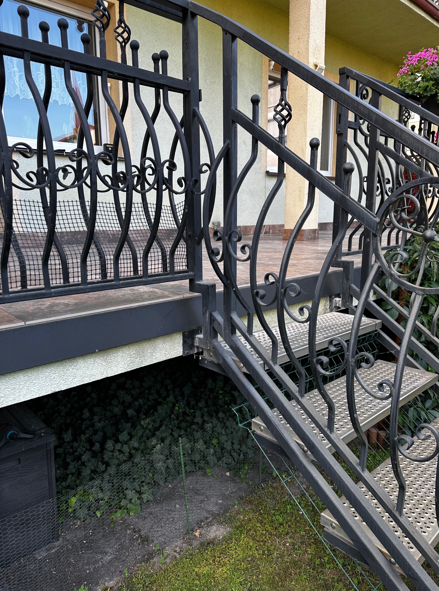 Metalowe schody zewnętrzne z ozdobnymi elementami prowadzące na taras domu. Widoczne detale konstrukcji oraz roślinność w tle.