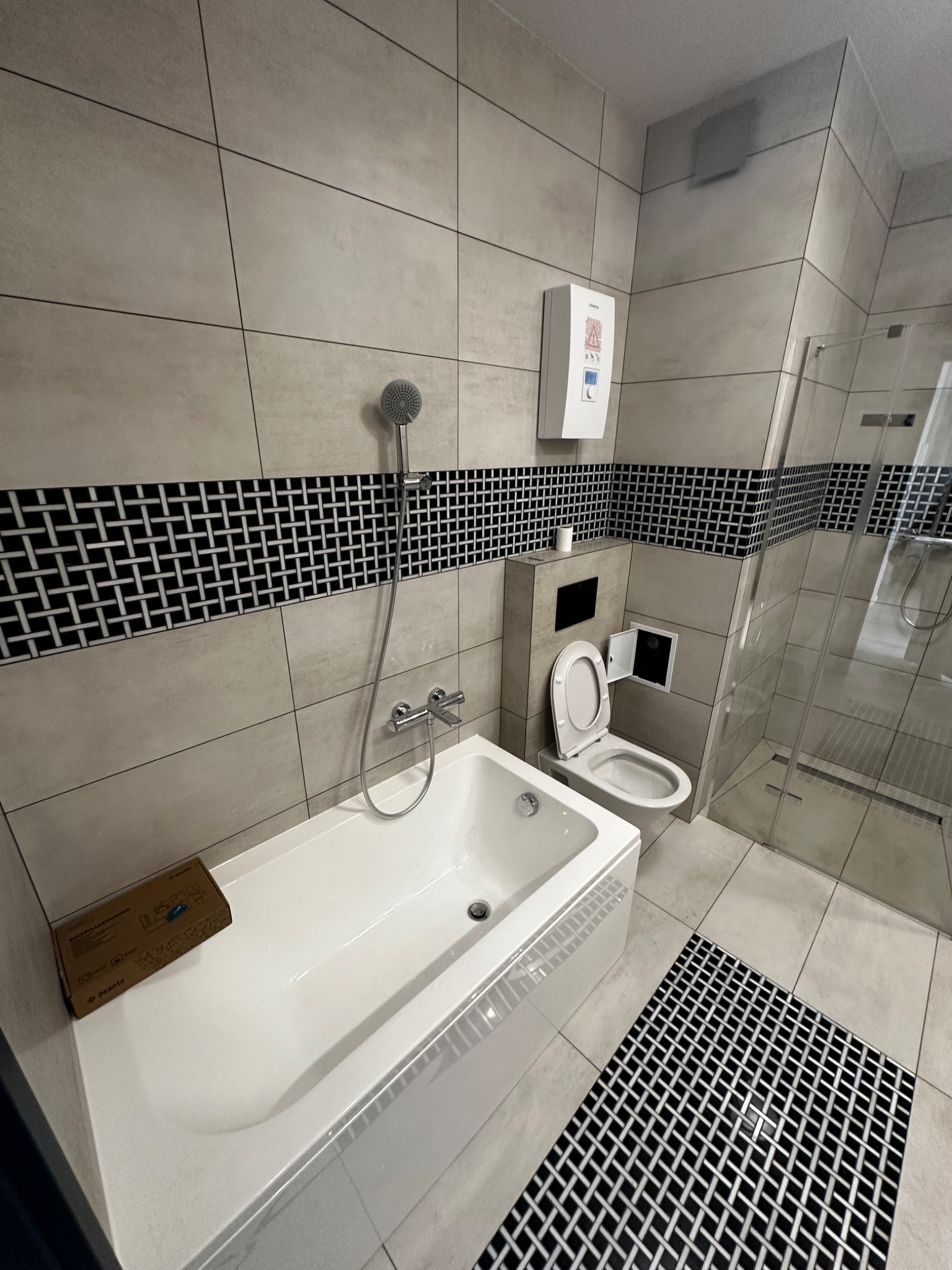 Jasna łazienka po renowacji z wanną, kabiną prysznicową i toaletą. Uwagę przyciąga dekoracyjny pas płytek w czarno-białą kratkę.