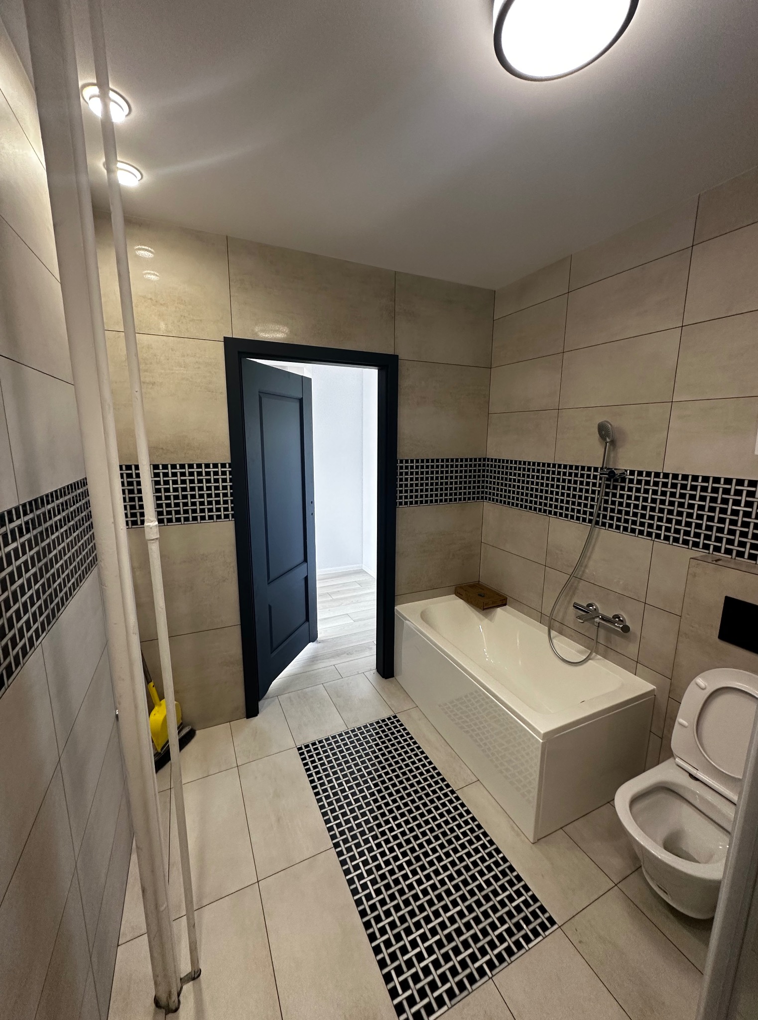 Wykończona łazienka z beżowymi płytkami, czarnym pasem dekoracyjnym, białą wanną i toaletą. Na podłodze dywanik w czarno-białą kratę. Czarne drzwi prowadzą do innego pomieszczenia.