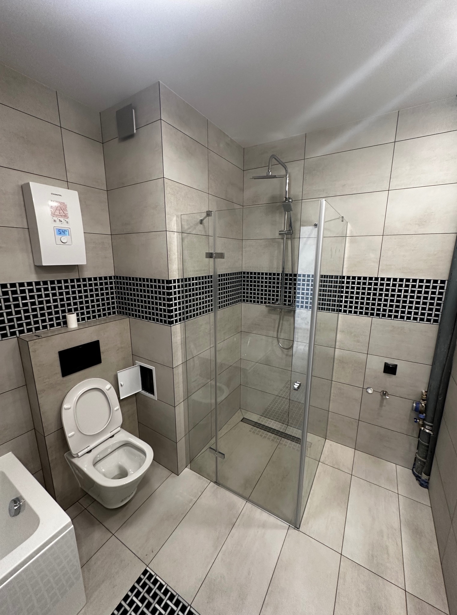 Wykończona łazienka w beżowych płytkach z czarnym pasem dekoracyjnym. Widoczna kabina prysznicowa ze szklanymi drzwiami, toaleta i fragment wanny. Oświetlenie sufitowe.