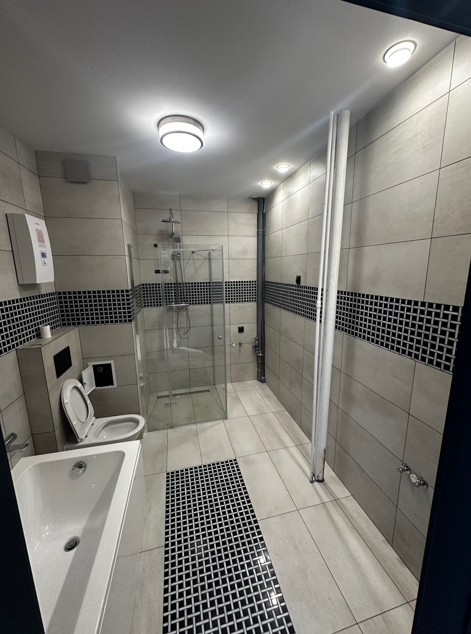 Wykończona łazienka w szarościach z kabiną prysznicową, toaletą i umywalką. Uwagę przykuwają czarno-białe, dekoracyjne pasy płytek oraz wzór na podłodze.