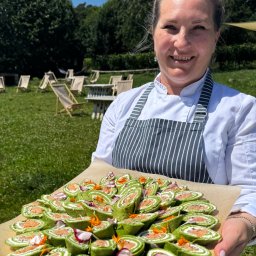 Restauracja Zagroda Jutrzenki 5 - Uśmiechnięta kucharka prezentuje tacę z zielonymi roladkami z łososiem na tle plenerowej imprezy z leżakami i stolikami. Słoneczny dzień.