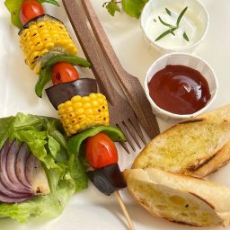 Restauracja Zagroda Jutrzenki 5 - Szpadka z grillowanymi warzywami (kukurydza, pomidor, bakłażan, papryka) na talerzu z pieczywem, sałatą i sosami. Ujęcie z góry.