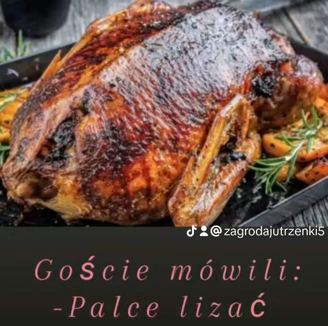 Apetyczna, pieczona kaczka z chrupiącą skórką, podana na czarnym talerzu z pieczonymi owocami i rozmarynem. Całość prezentuje się smakowicie i świątecznie.