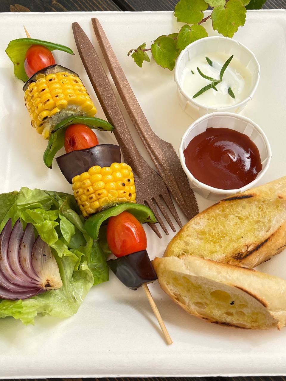 Szpadka z grillowanymi warzywami (kukurydza, pomidor, bakłażan, papryka) na talerzu z pieczywem, sałatą i sosami. Ujęcie z góry.