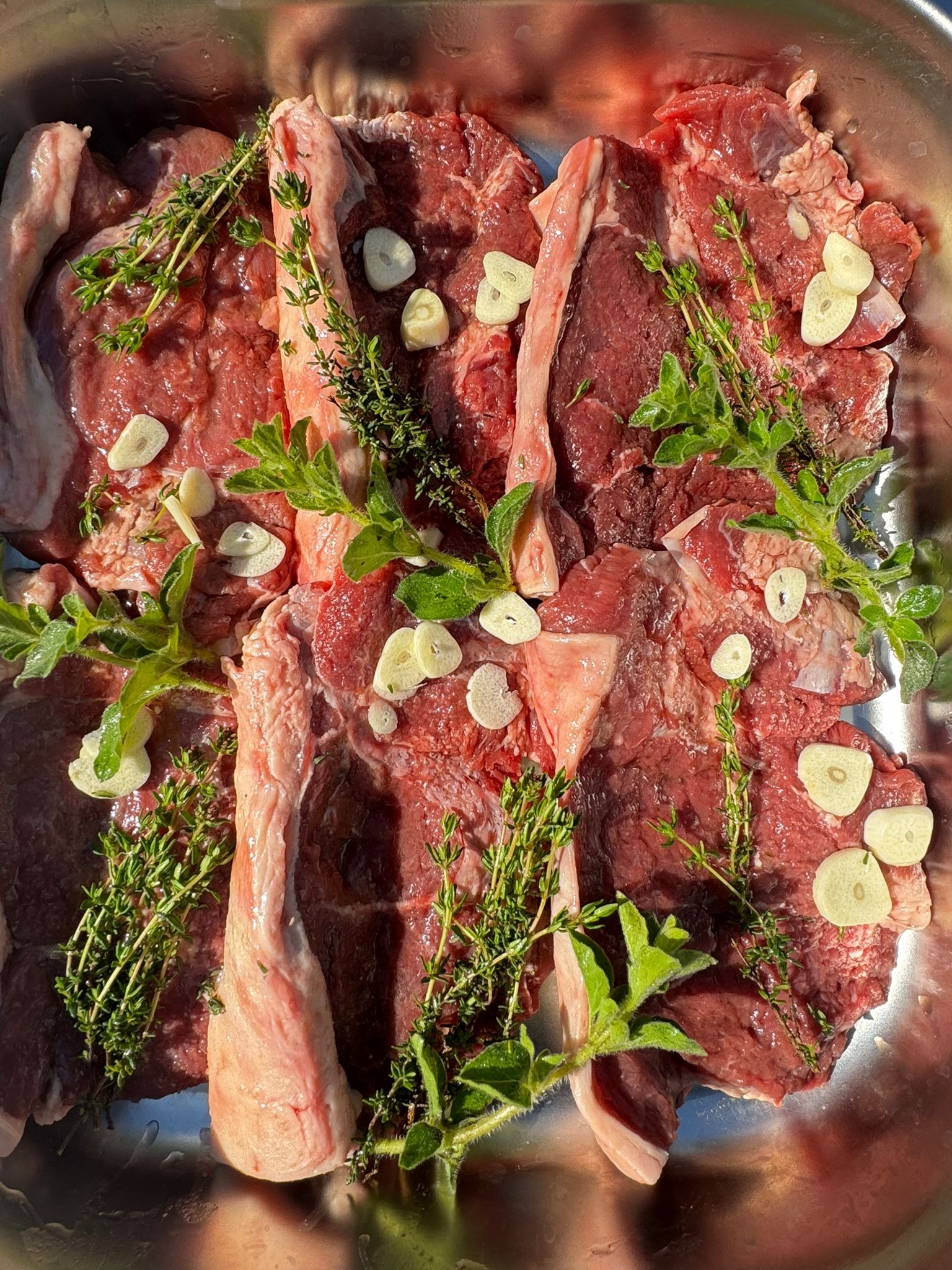 Surowe steki wołowe z kością, doprawione świeżym tymiankiem i czosnkiem w metalowej brytfannie, przygotowane do pieczenia lub grillowania.