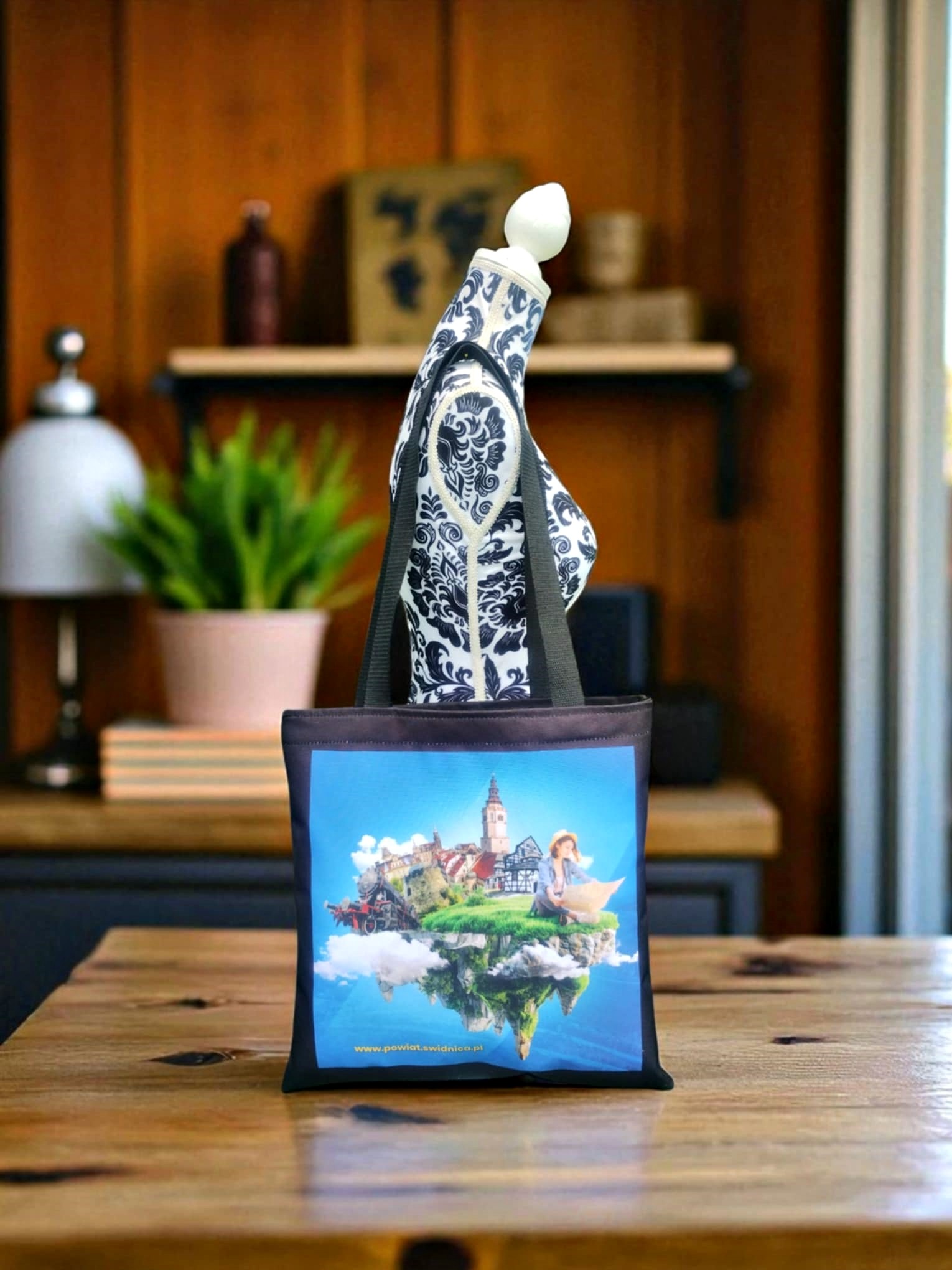 Torba bawełniana z nadrukiem krajobrazu, prezentowana na manekinie w studio. Widoczny adres strony internetowej na dole torby. Ciepłe oświetlenie, drewniany stół.