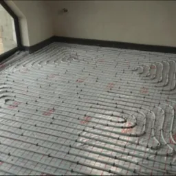 Białe rury ogrzewania podłogowego ułożone spiralnie na folii z czerwoną kratką w jasnym pomieszczeniu z oknem, widok z góry, etap instalacji przed wylaniem posadzki.