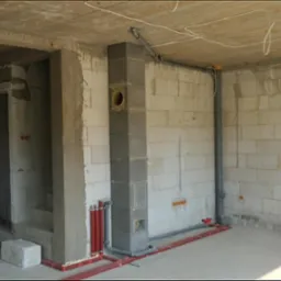 Wnętrze w trakcie instalacji: czerwone rury grzewcze przy ścianie z betonu komórkowego, szary komin wentylacyjny, pionowe rury kanalizacyjne na ścianie.