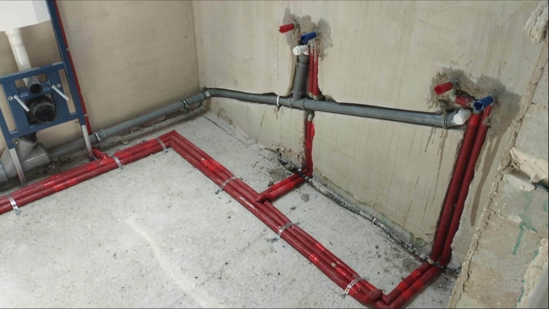 Nowa instalacja CO z czerwonymi rurami i szarą kanalizacją w surowym wnętrzu łazienki, przed położeniem wylewki i wykończeniem ścian.