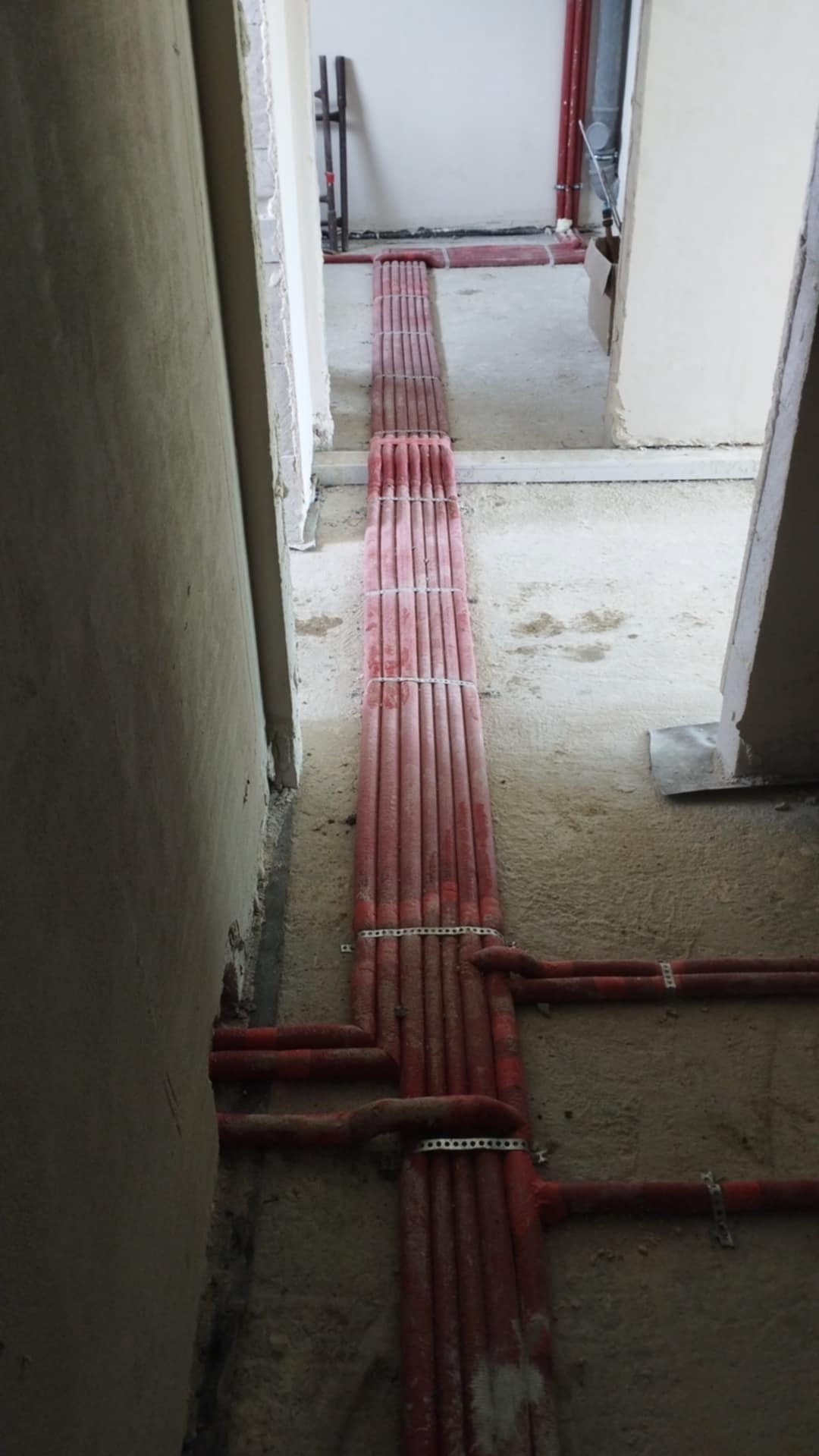 Czerwone rury instalacji grzewczej, ułożone równolegle na podłodze w surowym stanie, zabezpieczone białymi opaskami, widok z góry, w tle fragment ściany.