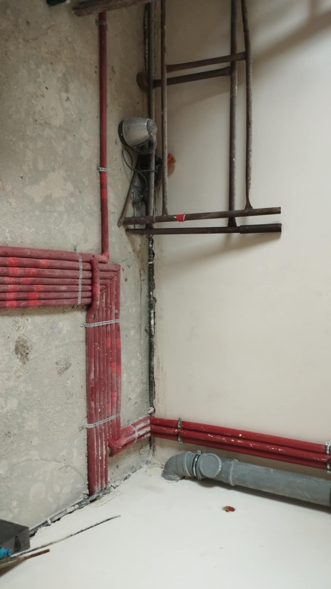 Czerwone rury instalacji grzewczej zamocowane na ścianie w surowym wnętrzu, widoczne szare rury kanalizacyjne przy podłodze i metalowa konstrukcja pod sufitem.