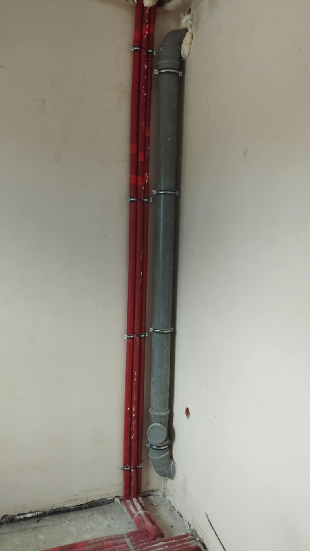 Czerwone rury CO oraz szara rura kanalizacyjna zamocowane do ściany klamrami. Rury w rogu pomieszczenia, widoczne uszczelnienia pianką montażową.