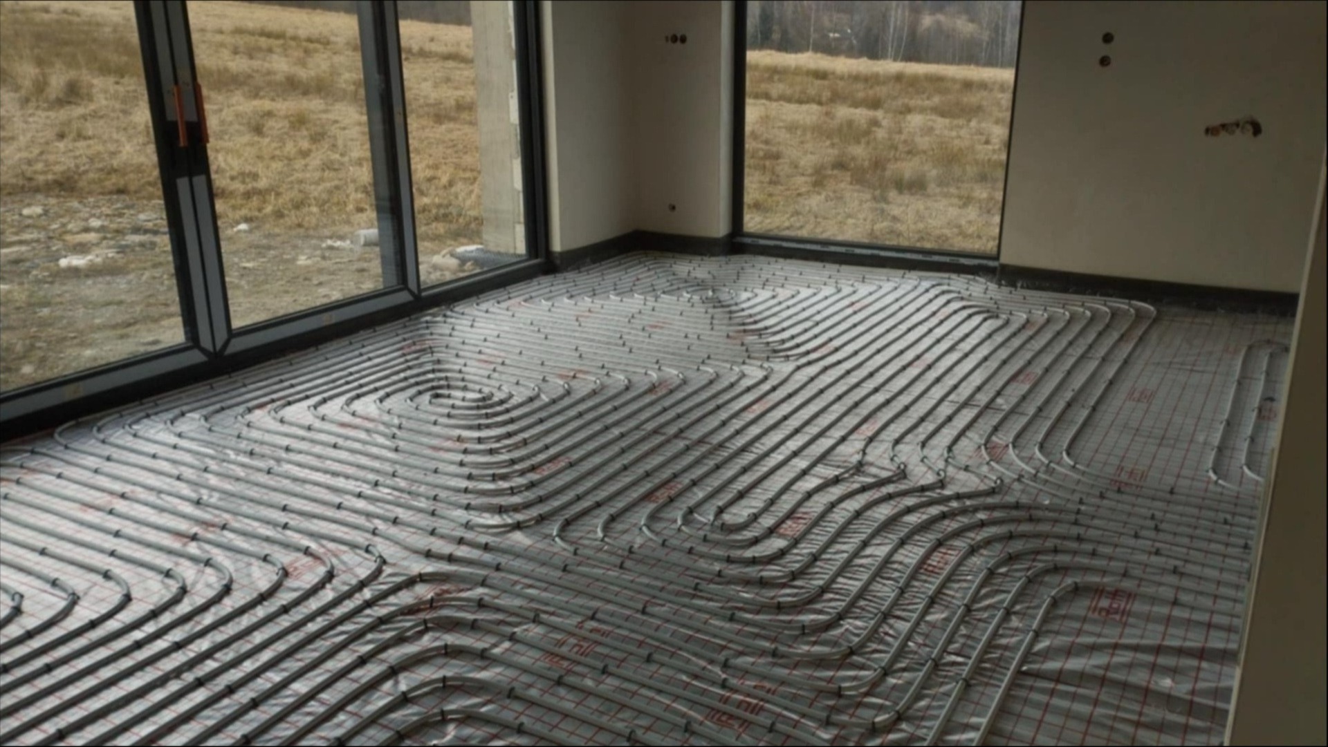 Instalacja ogrzewania podłogowego w nowym domu z dużymi oknami. Szare rury ułożone na folii z czerwoną siatką, widok na zewnątrz przez okno.