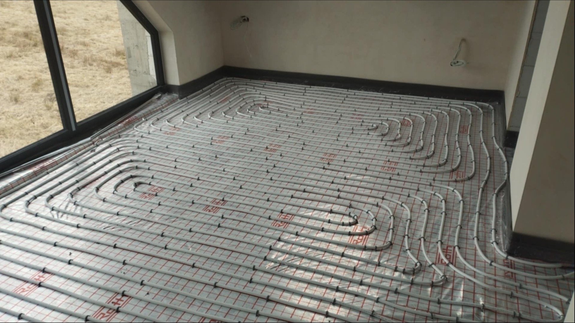 Białe rury ogrzewania podłogowego ułożone spiralnie na folii z czerwoną kratką w jasnym pomieszczeniu z oknem, widok z góry, etap instalacji przed wylaniem posadzki.