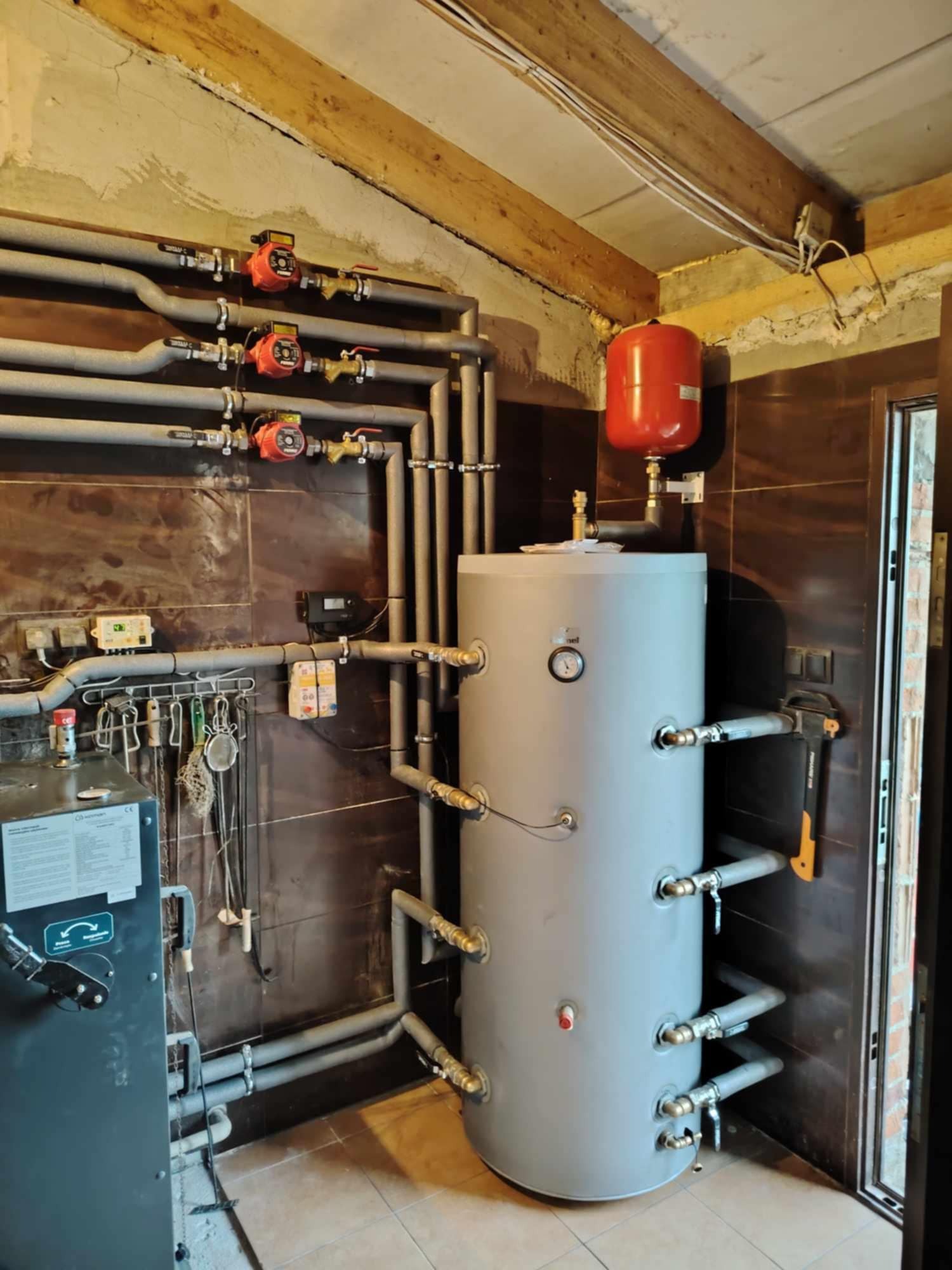 Złożona instalacja grzewcza z szarym zbiornikiem akumulacyjnym, czerwonym naczyniem wzbiorczym i licznymi rurami oraz pompami obiegowymi w kotłowni z brązowymi płytkami.