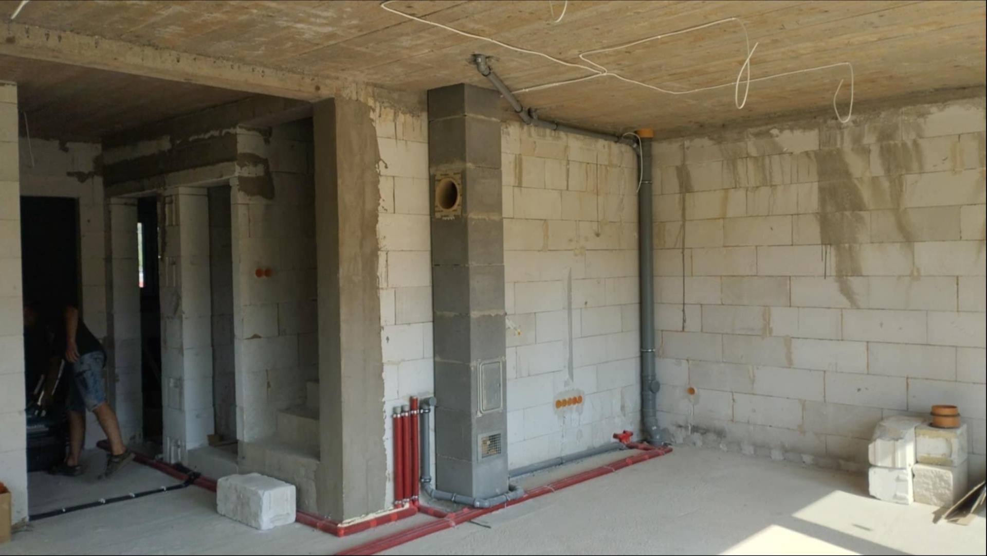 Wnętrze w trakcie instalacji: czerwone rury grzewcze przy ścianie z betonu komórkowego, szary komin wentylacyjny, pionowe rury kanalizacyjne na ścianie.