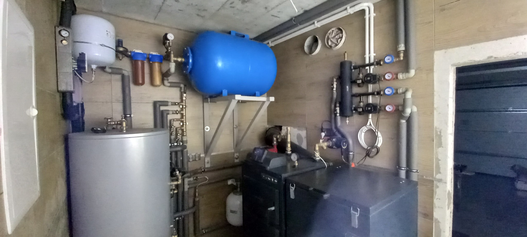 Kompletna kotłownia z szarym bojlerem, niebieskim zbiornikiem hydroforowym i filtrami wody. Instalacja z rur i zaworów na tle drewnopodobnych płytek.