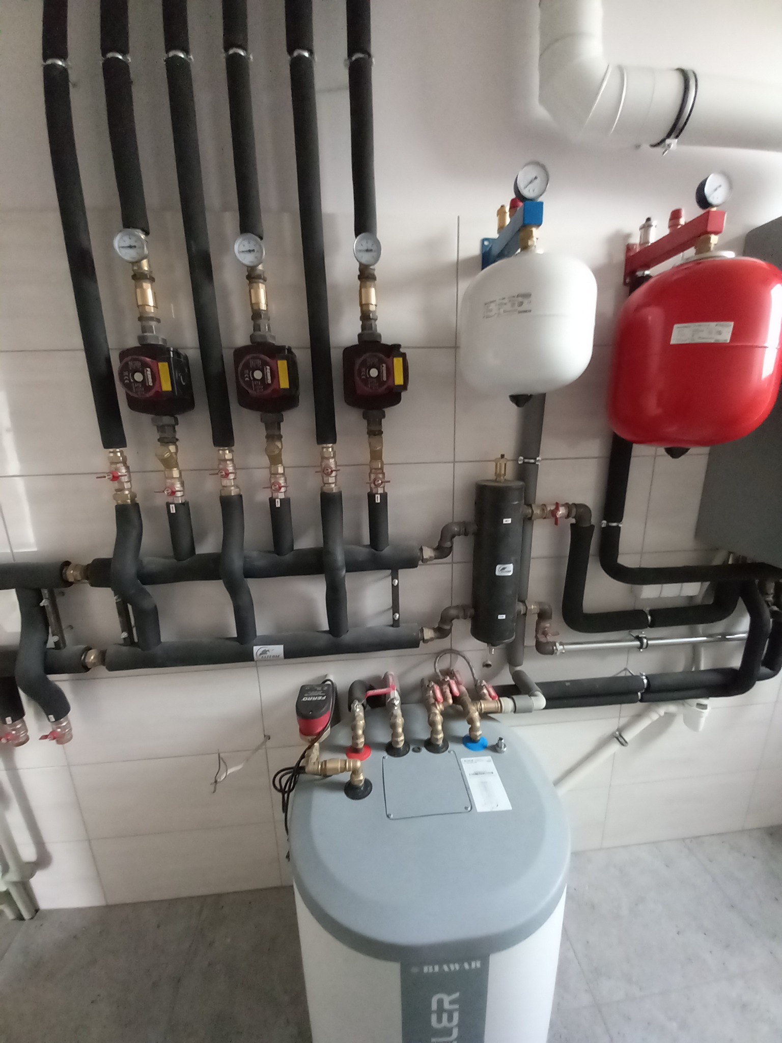 Węzeł cieplny z pompami obiegowymi, zbiornikiem i orurowaniem, manometry na rurach w izolacji termicznej, biało-szare płytki na ścianach i podłodze.