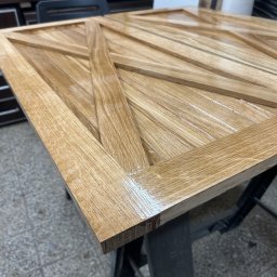 BAYWOOD - usługi stolarskie i ślusarskie - Lakierowany, drewniany panel z geometrycznym wzorem, ustawiony na metalowym stojaku. Widoczne detale struktury drewna i połysk lakieru. Warsztat stolarski w tle.