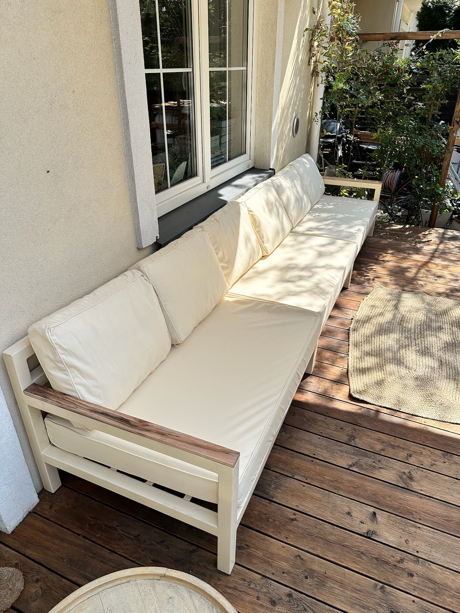 Długi, kremowy narożnik z jasnymi poduchami na drewnianym tarasie. Widoczne drewniane deski tarasowe i fragment dywanu. W tle budynek z oknami i roślinność.
