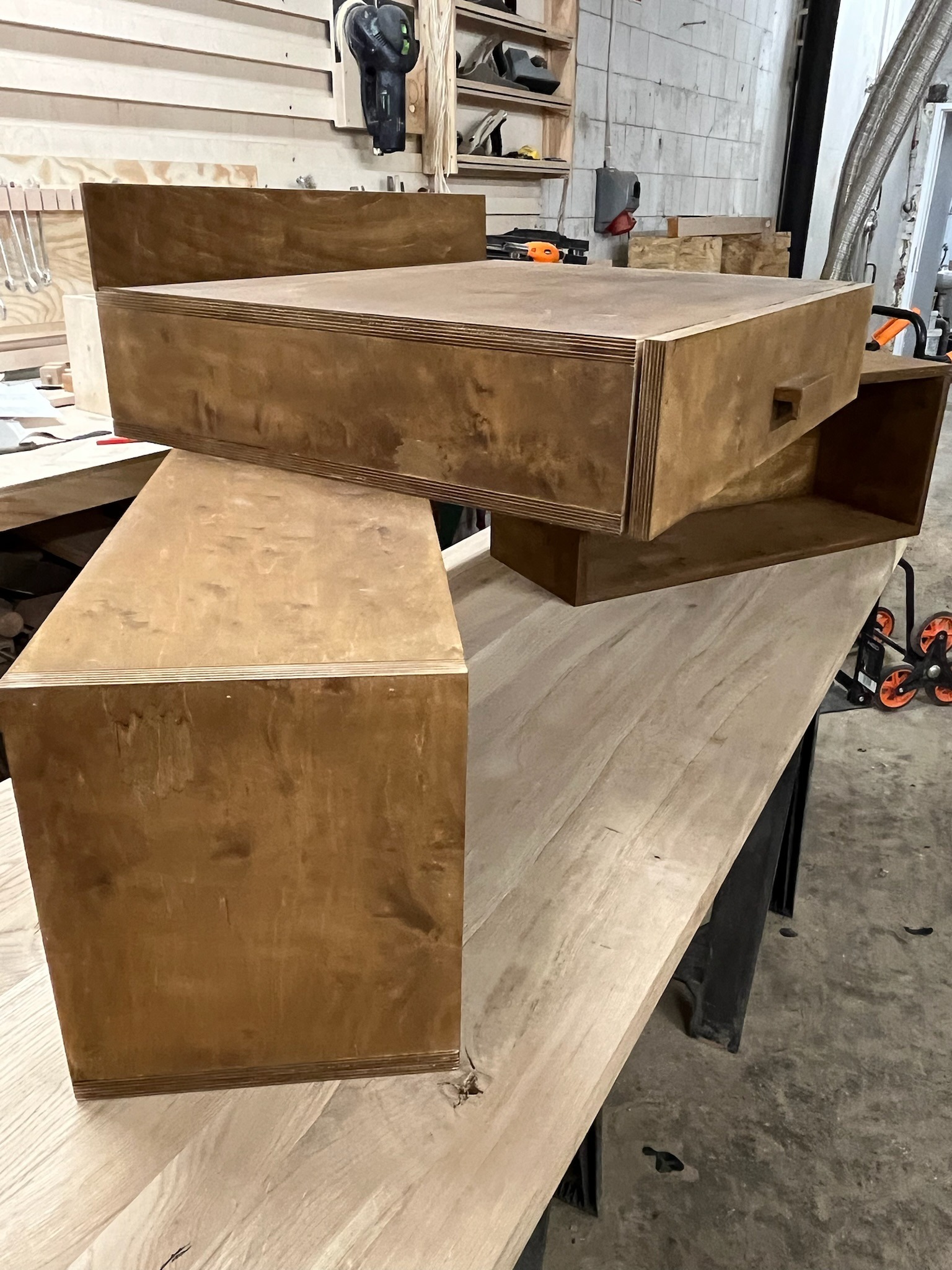 Brązowe, drewniane elementy meblowe z widocznymi warstwami sklejki, ułożone na blacie w warsztacie. Elementy o różnych kształtach, prawdopodobnie przygotowane do montażu.