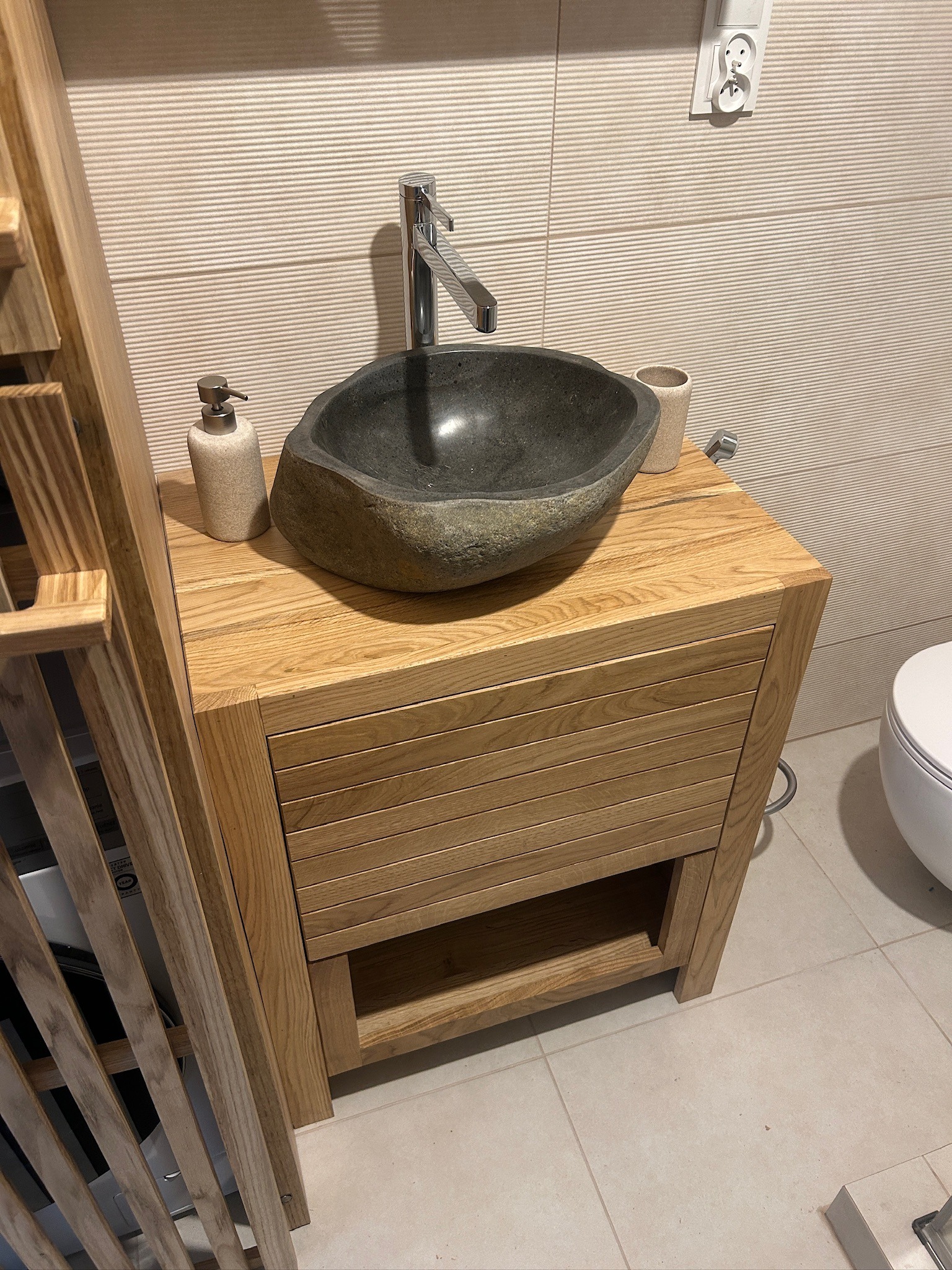 Unikalna, kamienna umywalka osadzona na drewnianej szafce w łazience. Widoczny dozownik mydła i fragment toalety. Tło stanowią płytki imitujące drewno.