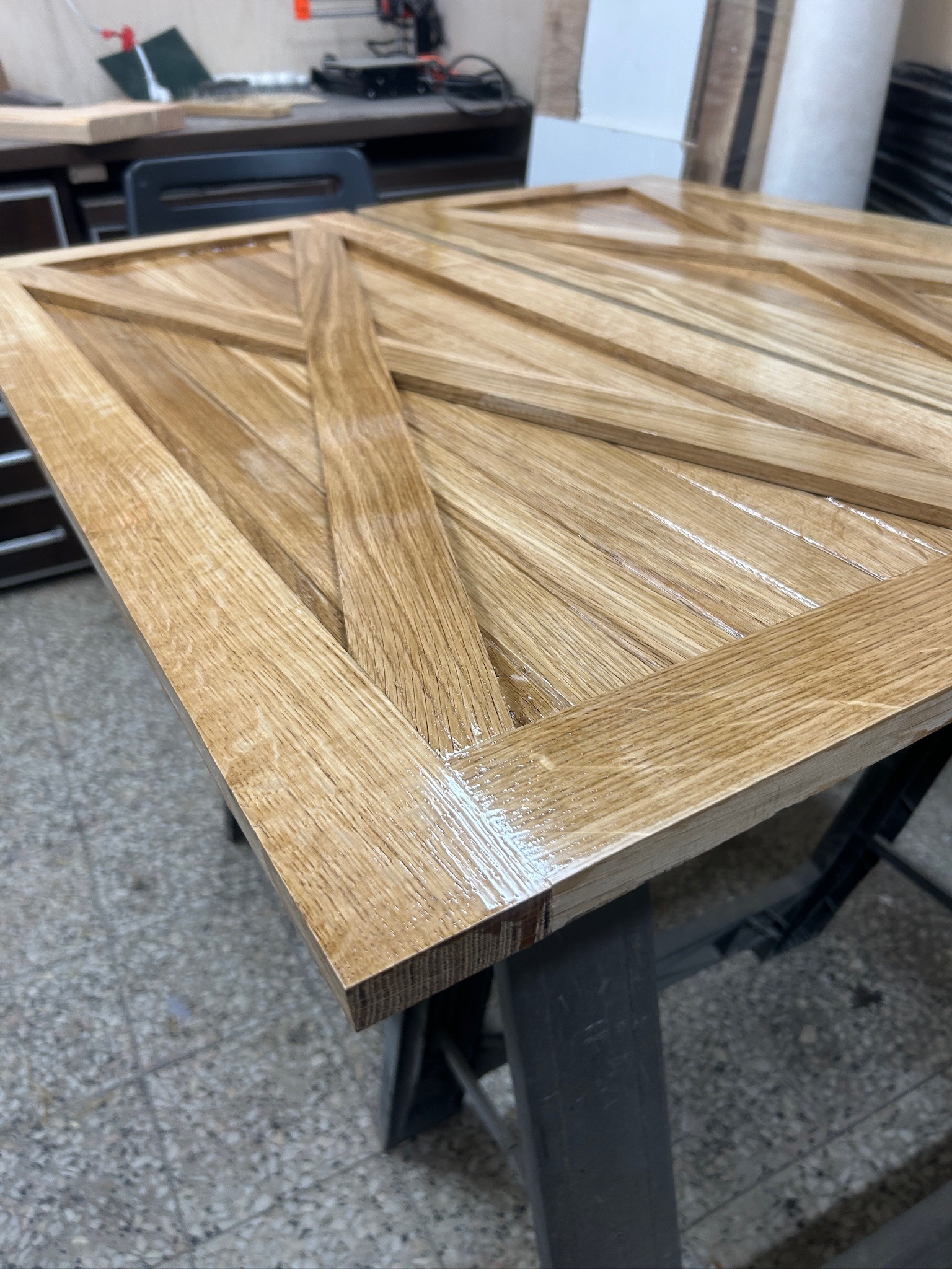 Lakierowany, drewniany panel z geometrycznym wzorem, ustawiony na metalowym stojaku. Widoczne detale struktury drewna i połysk lakieru. Warsztat stolarski w tle.