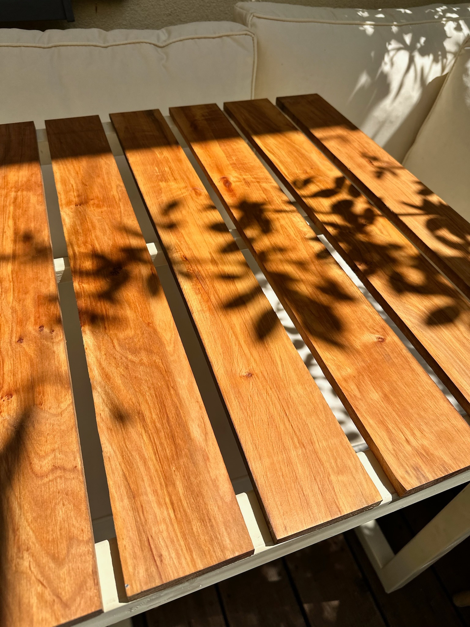 Drewniane deski z widoczną strukturą drewna, ułożone równolegle na białej konstrukcji, oświetlone słońcem i cieniami liści. Widok z góry, bliski plan.