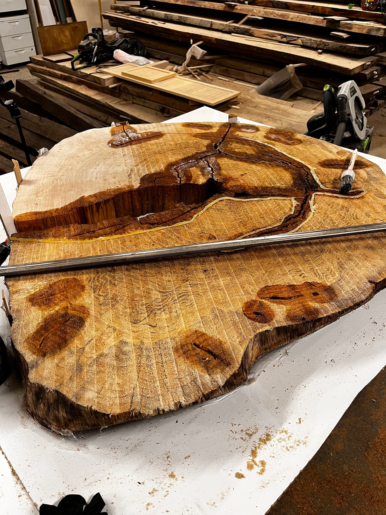 Obrabiany plaster drewna z widocznymi żywicznymi wypełnieniami, zabezpieczony metalową listwą. W tle warsztat z narzędziami i deskami.